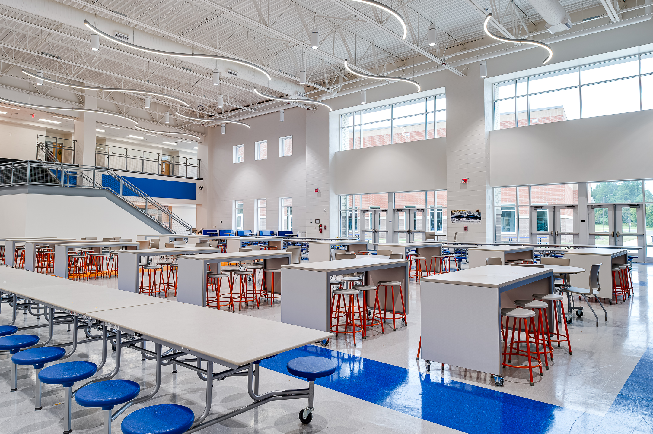 A high school cafeteria with big windows, orange stools and blue benches