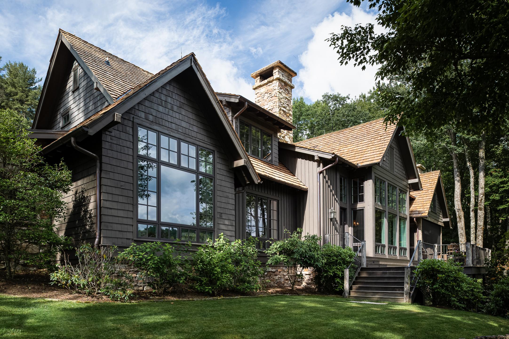 A modern, dark-colored house with large windows and a stone chimney is surrounded by trees and has a well-maintained lawn, beautifully captured by a North Carolina architectural photographer.