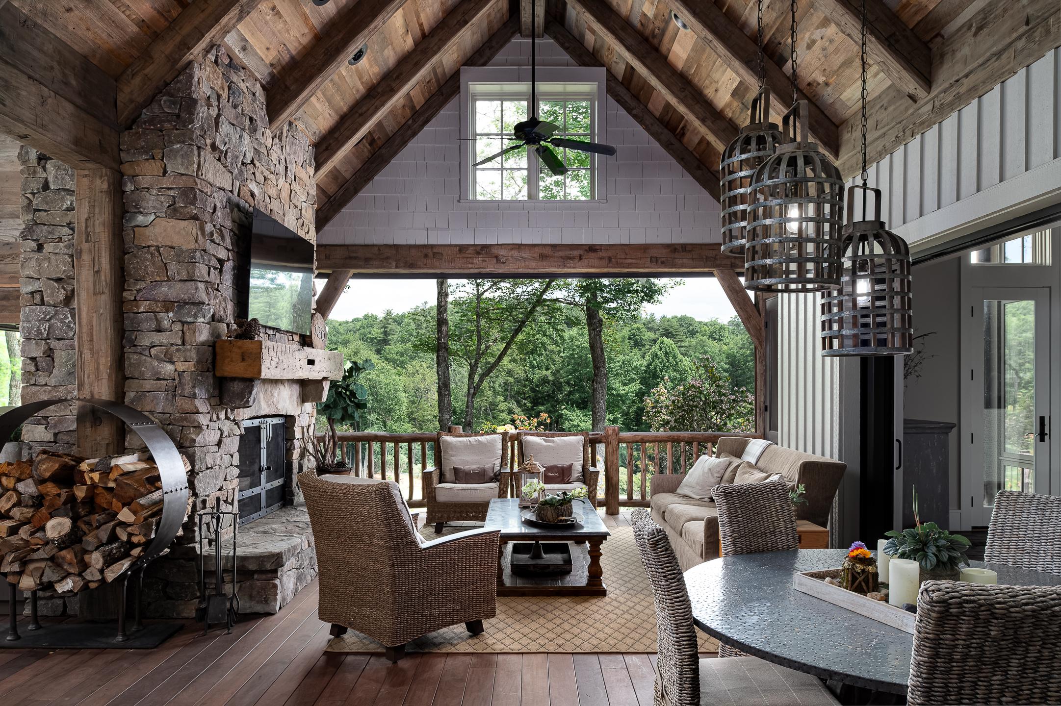 A rustic outdoor living space with wooden beams and a stone fireplace, furnished with wicker chairs, a coffee table, and a dining area, overlooks a lush green landscape through a large open window. Perfect for an architectural photographer capturing the harmony between nature and design. captured by professional architectural photographer