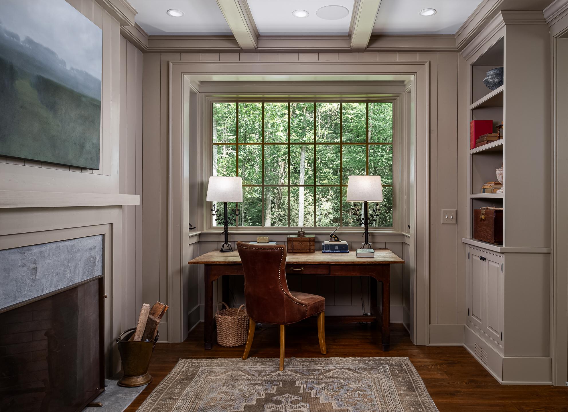 A home office with a wooden desk, a leather chair, two lamps, and built-in shelves. A large window with a view of North Carolina greenery is behind the desk. The room features paneled walls, a framed landscape painting, and is perfect for an architectural photographer seeking inspiration.