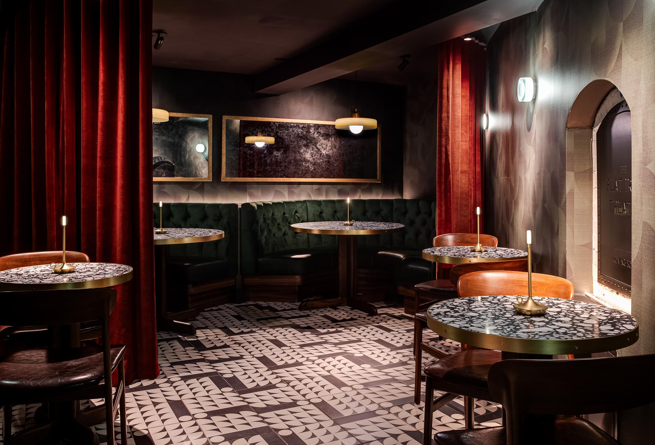 A dimly lit restaurant interior with round tables, mosaic patterned floors, red curtains, and cushioned green seating. Wall mirrors reflect the modern decor, captured perfectly by an architectural photographer. Small lamps are on each table.